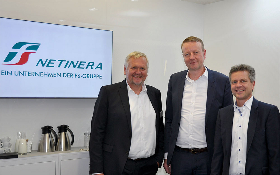 Group picture against a white background. On the left is a screen with the logo of NETINERA, below it a small sideboard with glasses, cups and coffee pots.