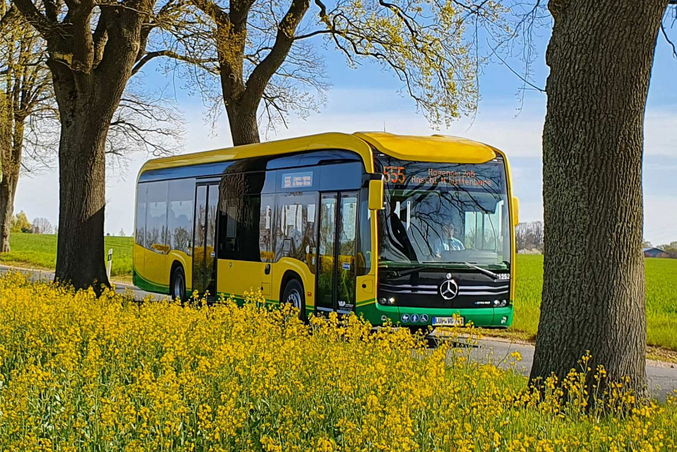 A yellow VLP electric bus on a road between yellow rapeseed fields