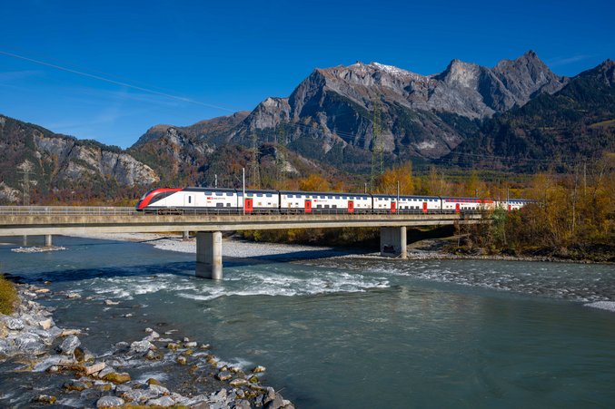An SBB train drives via a river in the Alps