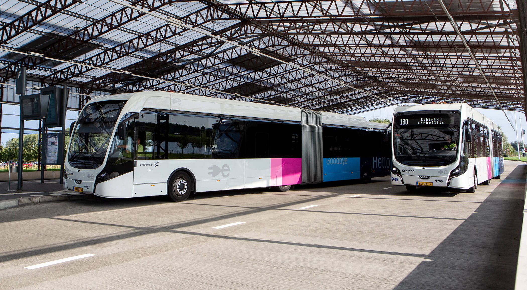 Two modern buses at a covered bus terminal with steel roof structure