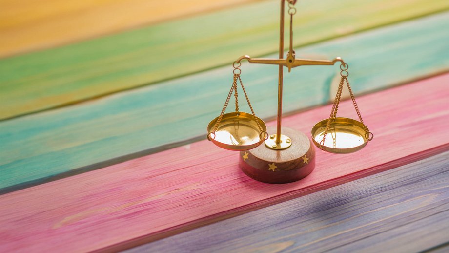 Eine kleine goldene Waage auf einem Holztisch dessen Holzleisten regenbogenfarben angemalt sind und für Chancengerechtigkeit steht