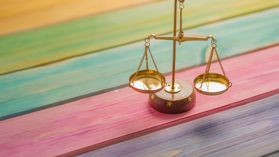A small golden scale on a wooden table whose wooden slats are painted in rainbow colors, symbolizing equal opportunity