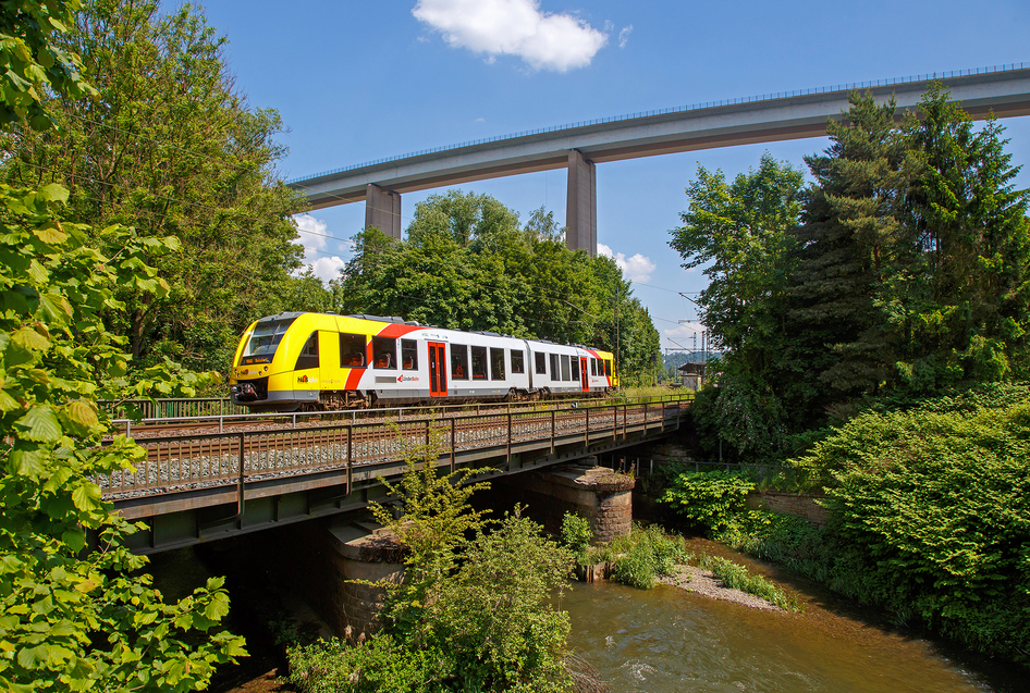 A white and yellow HLB train crosses a bridge that leads over a river in a forest.