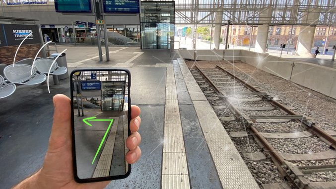 Hand hält Smartphone mit App-Anzeige auf leerem Bahnsteig mit Gleisen