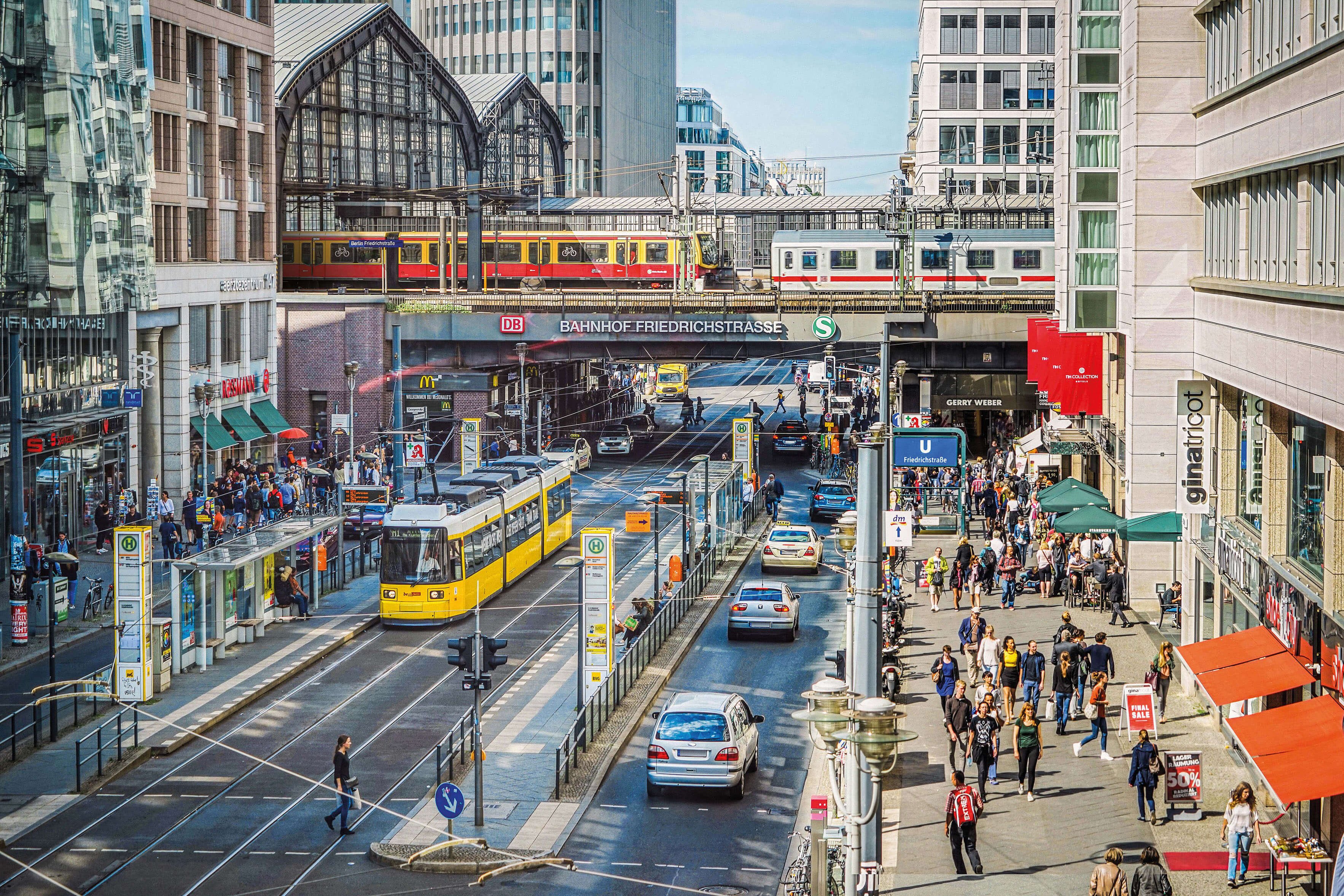 Concurrida Friedrichstraße en Berlín con S-Bahn, tranvía amarillo, peatones y la estación Friedrichstraße al fondo