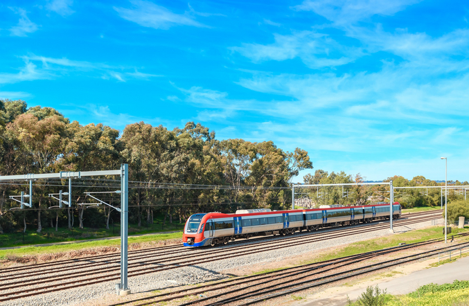 A red and silver Keolis train moves from right to left in front of trees and a bright blue sky.