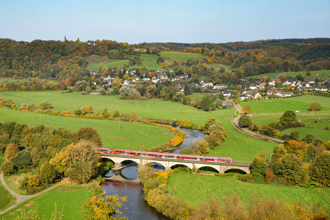 A red DB Regio train crosses a bridge over a river in the countryside with a small town in the background.