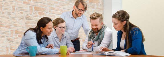 Fünf Kollegen sitzen an einem Tisch und diskutieren die darauf liegenden Dokumente