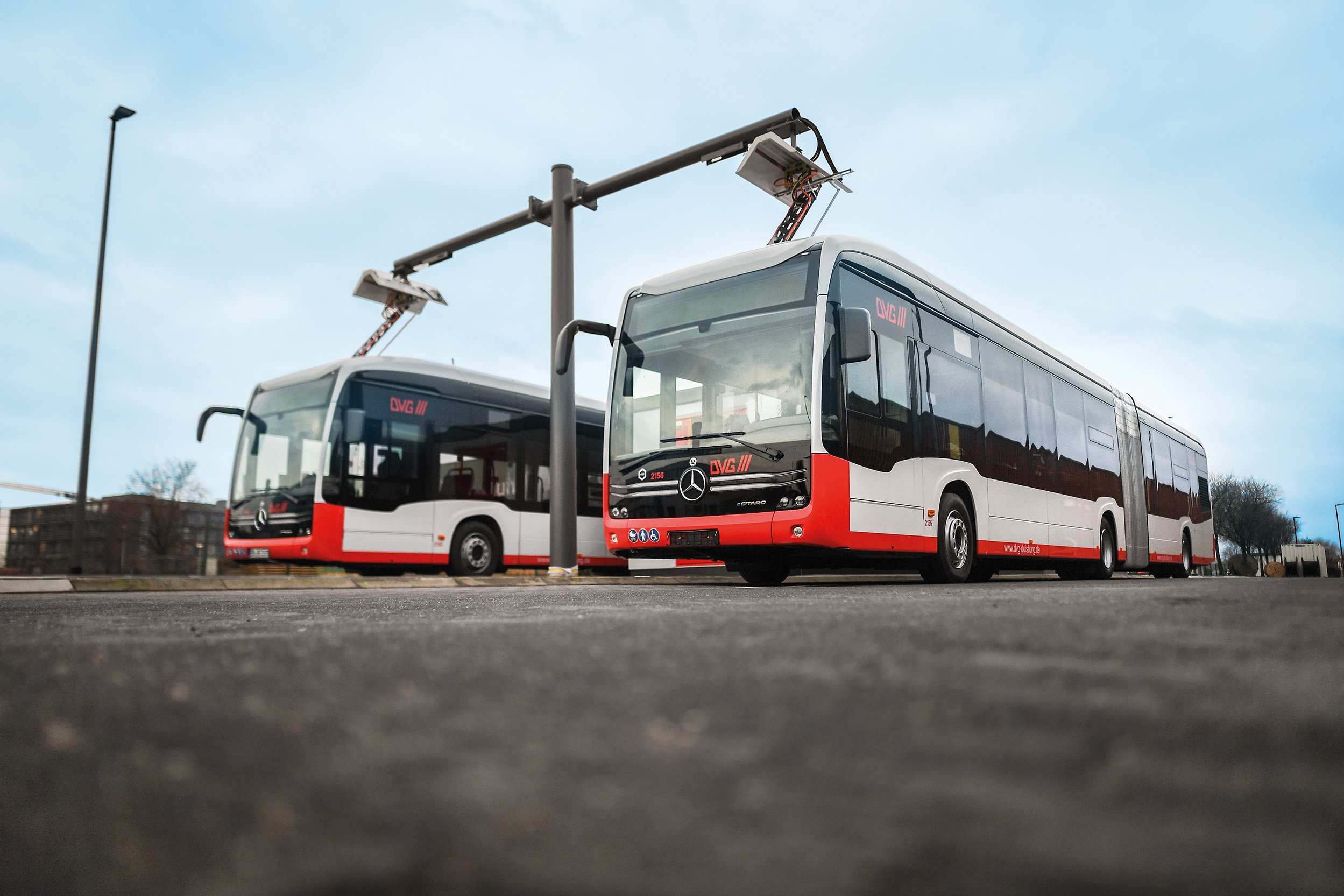 Two DVG electric buses in red and white livery charging at a depot charging station.