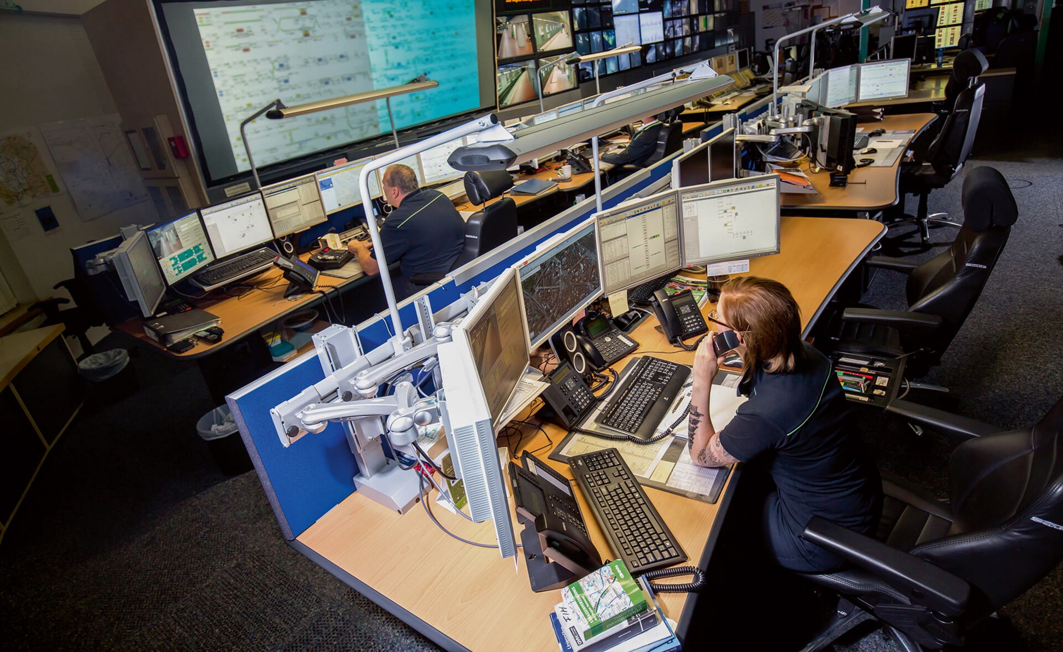 Operator at a workstation in a modern public transport control centre with multiple monitors and large display screen