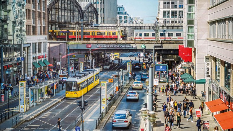Belebte Friedrichstraße in Berlin mit S-Bahn, gelber Straßenbahn, Fußgängern und dem Bahnhof Friedrichstraße im Hintergrund