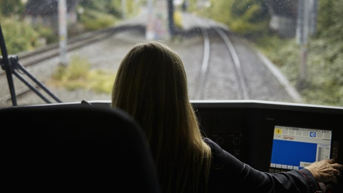 Woman in train cab looking at tracks through windscreen with laptop in foreground