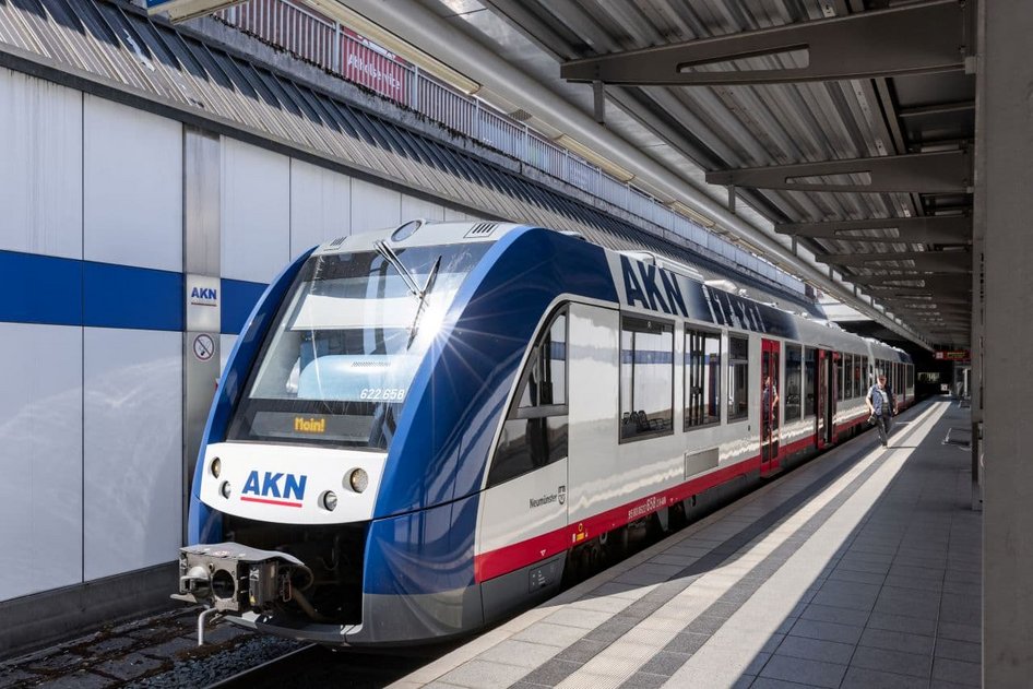Tren AKN en colores azul, blanco y rojo en andén de estación ferroviaria cubierta.