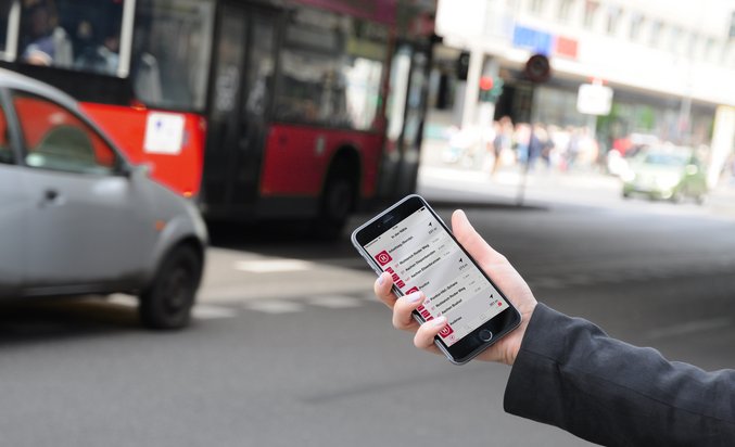 Eine Hand hält Handy mit Fahrgast-App ASEAG mobil an einer Straße mit Bus und Auto im Hintergrund in die Kamera