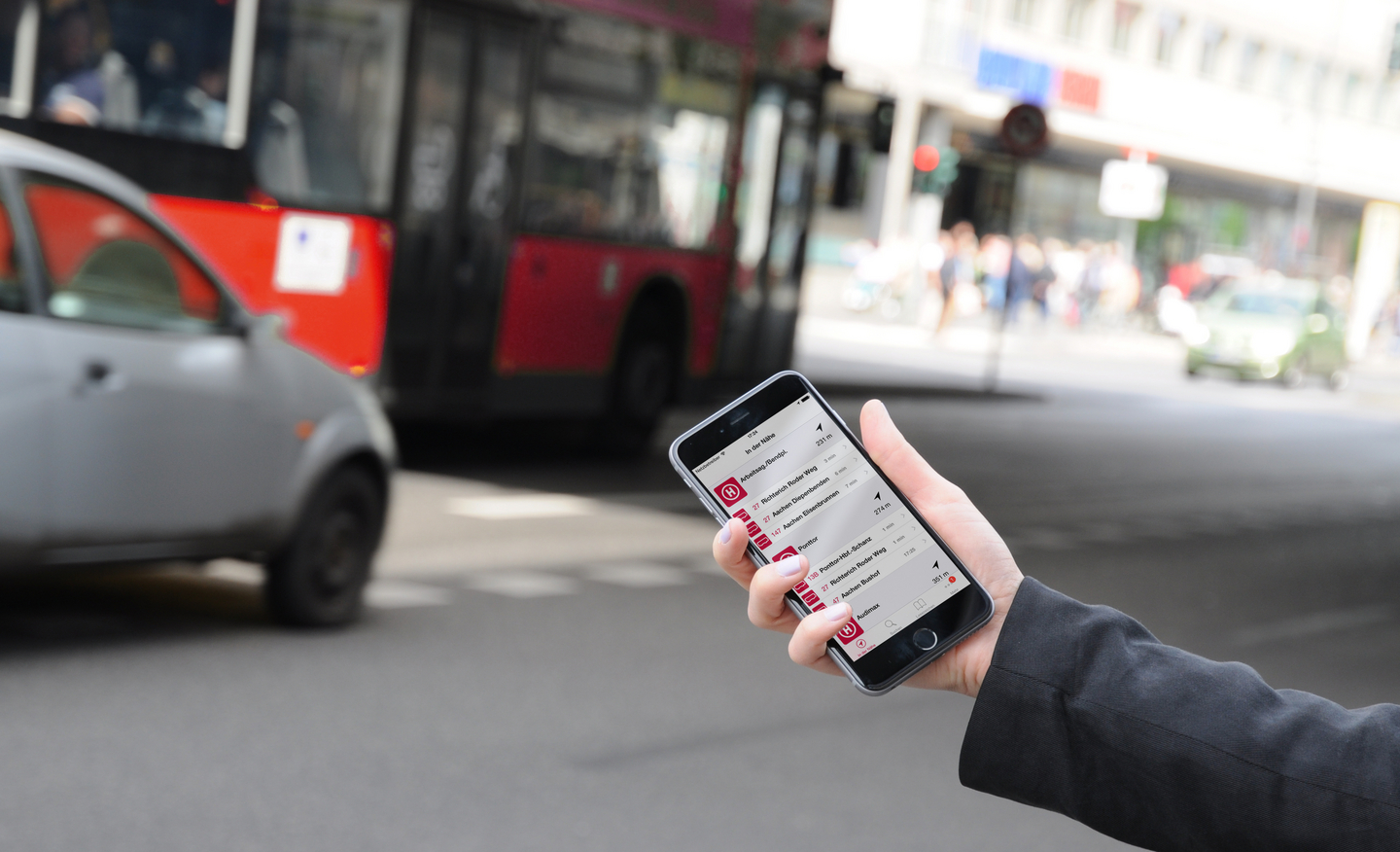 Eine Hand hält Handy mit Fahrgast-App ASEAG mobil an einer Straße mit Bus und Auto im Hintergrund in die Kamera