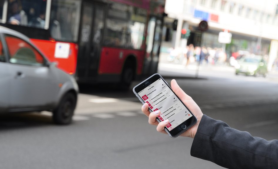Eine Hand hält Handy mit Fahrgast-App ASEAG mobil an einer Straße mit Bus und Auto im Hintergrund in die Kamera