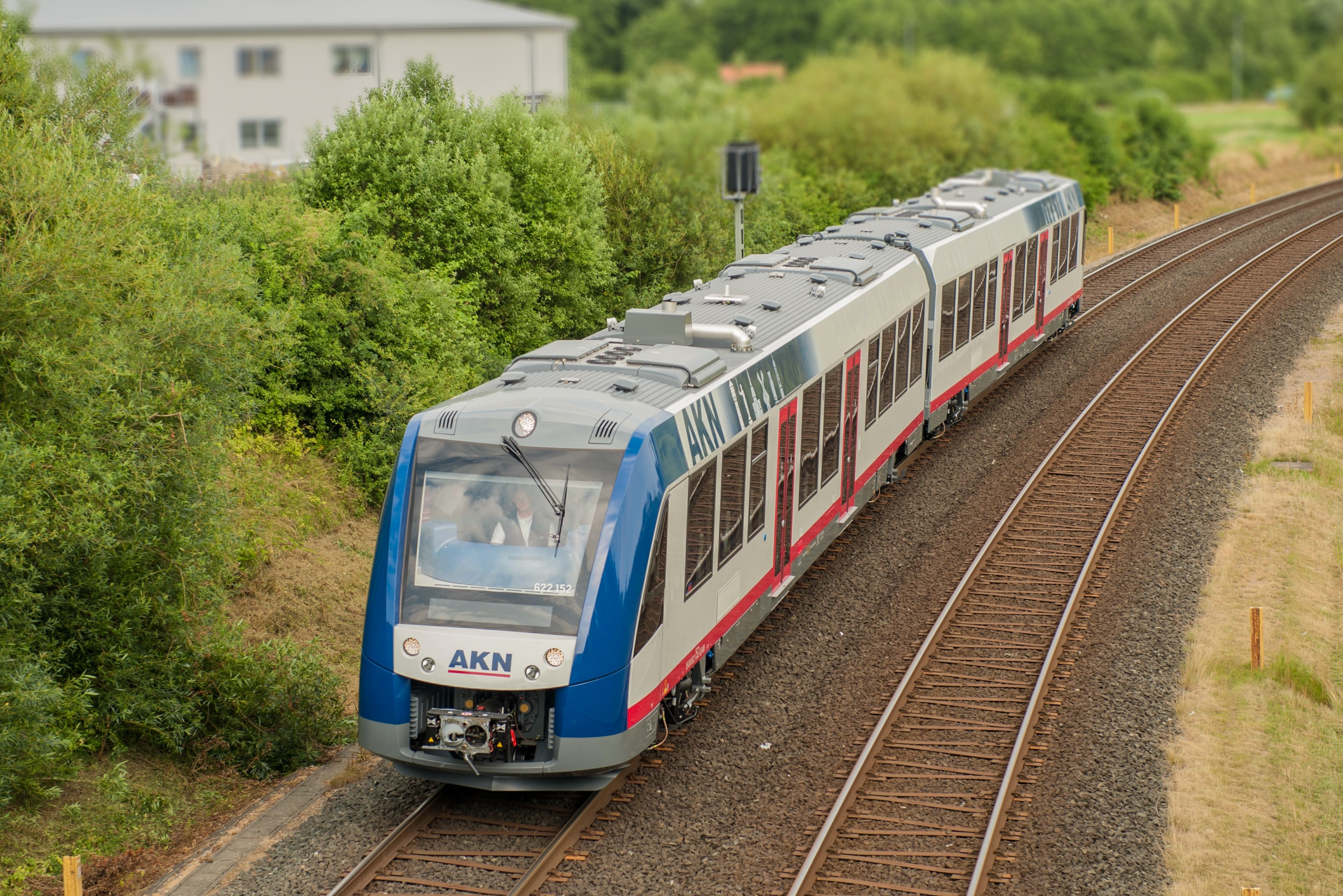Ein blau-weiß-roter AKN Zug fährt nahe einer Ortschaft im Grünen.