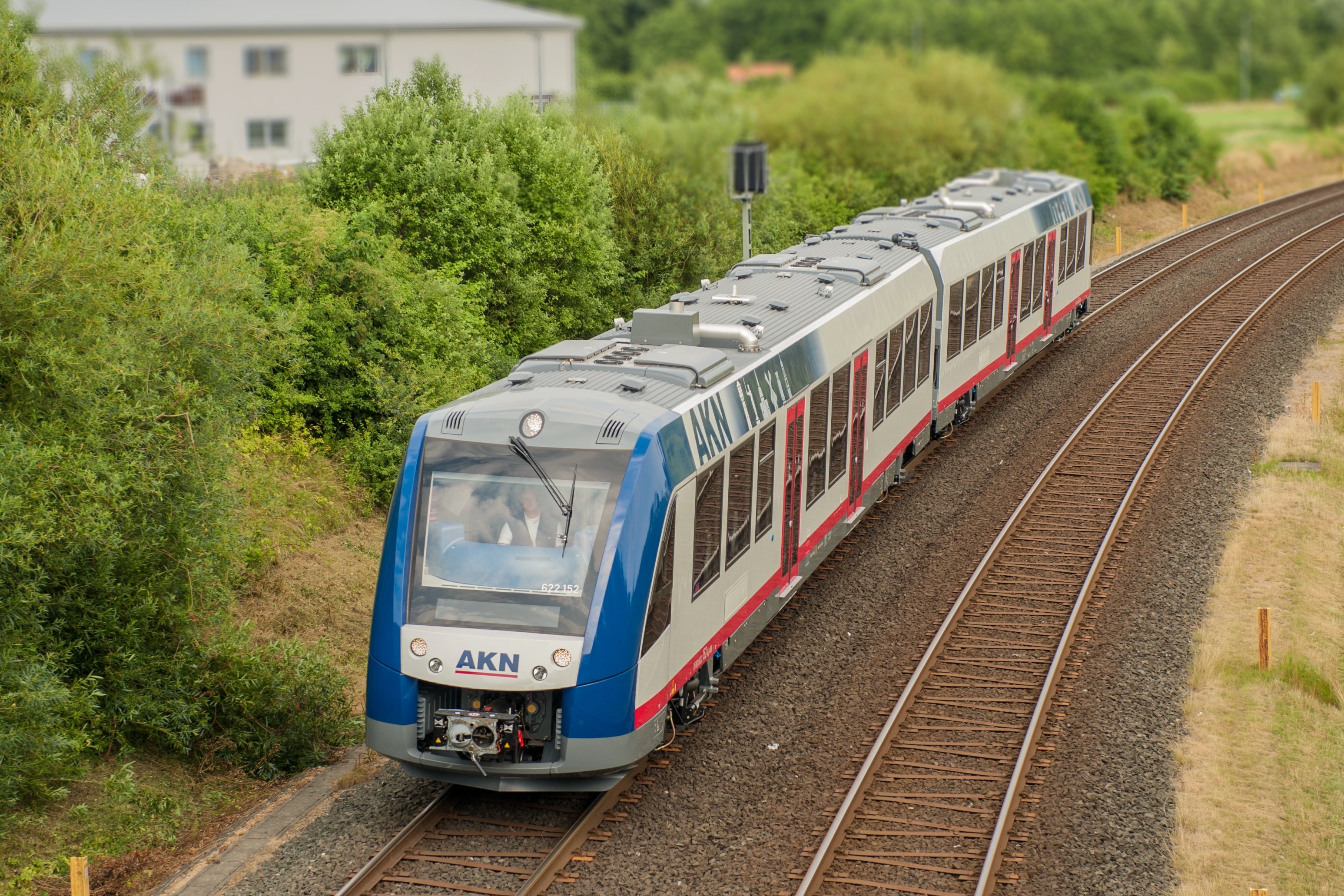 AKN railcar in blue, white and red livery running on single-track line through green countryside.