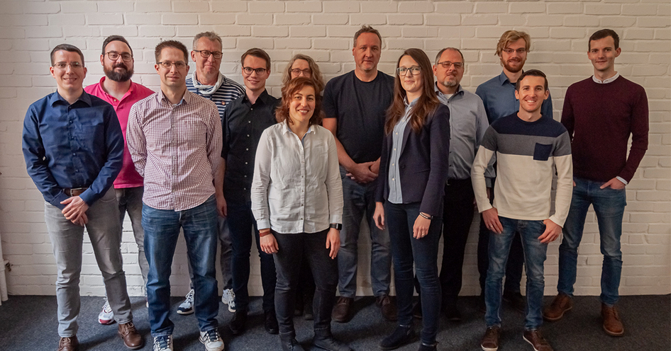 Gruppenfoto von dreizehn Menschen vor einer grauen Wand