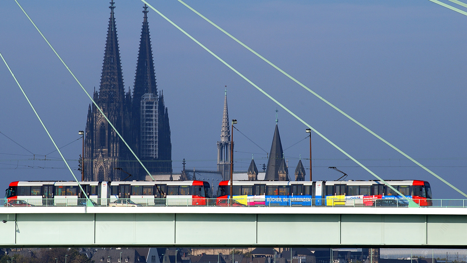 The Cologne tram runs on a bridge with Cologne Cathedral in the background.