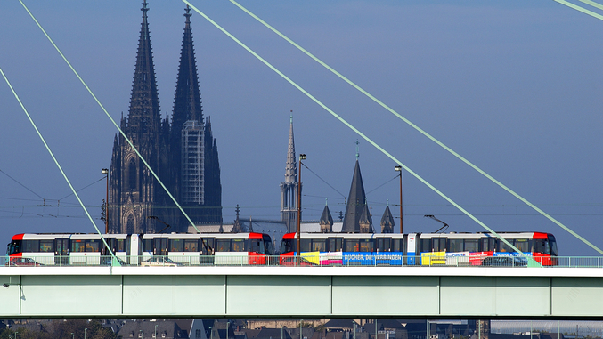 The Cologne tram runs on a bridge with Cologne Cathedral in the background.