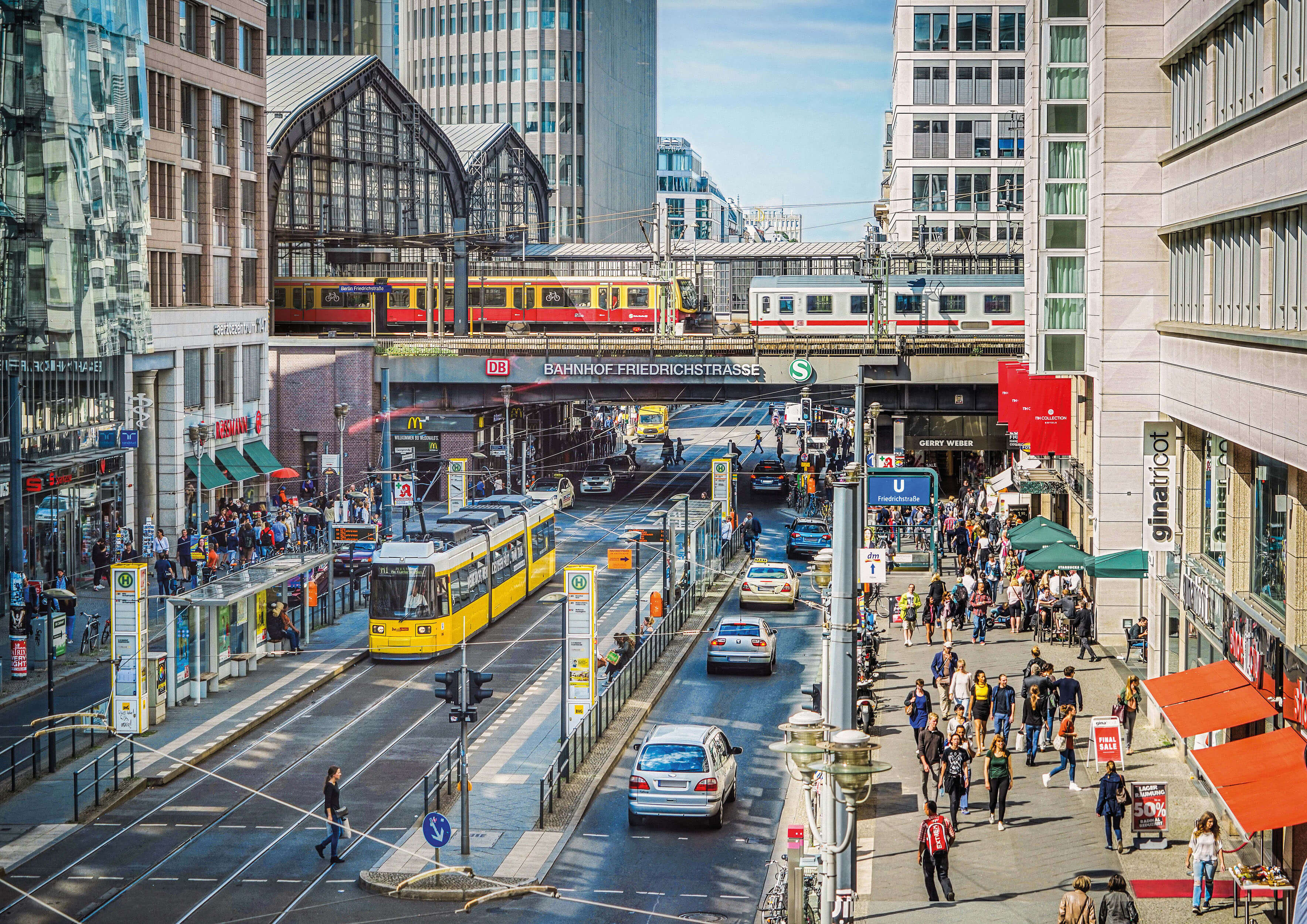 Belebte Friedrichstraße in Berlin mit S-Bahn, gelber Straßenbahn, Fußgängern und dem Bahnhof Friedrichstraße im Hintergrund