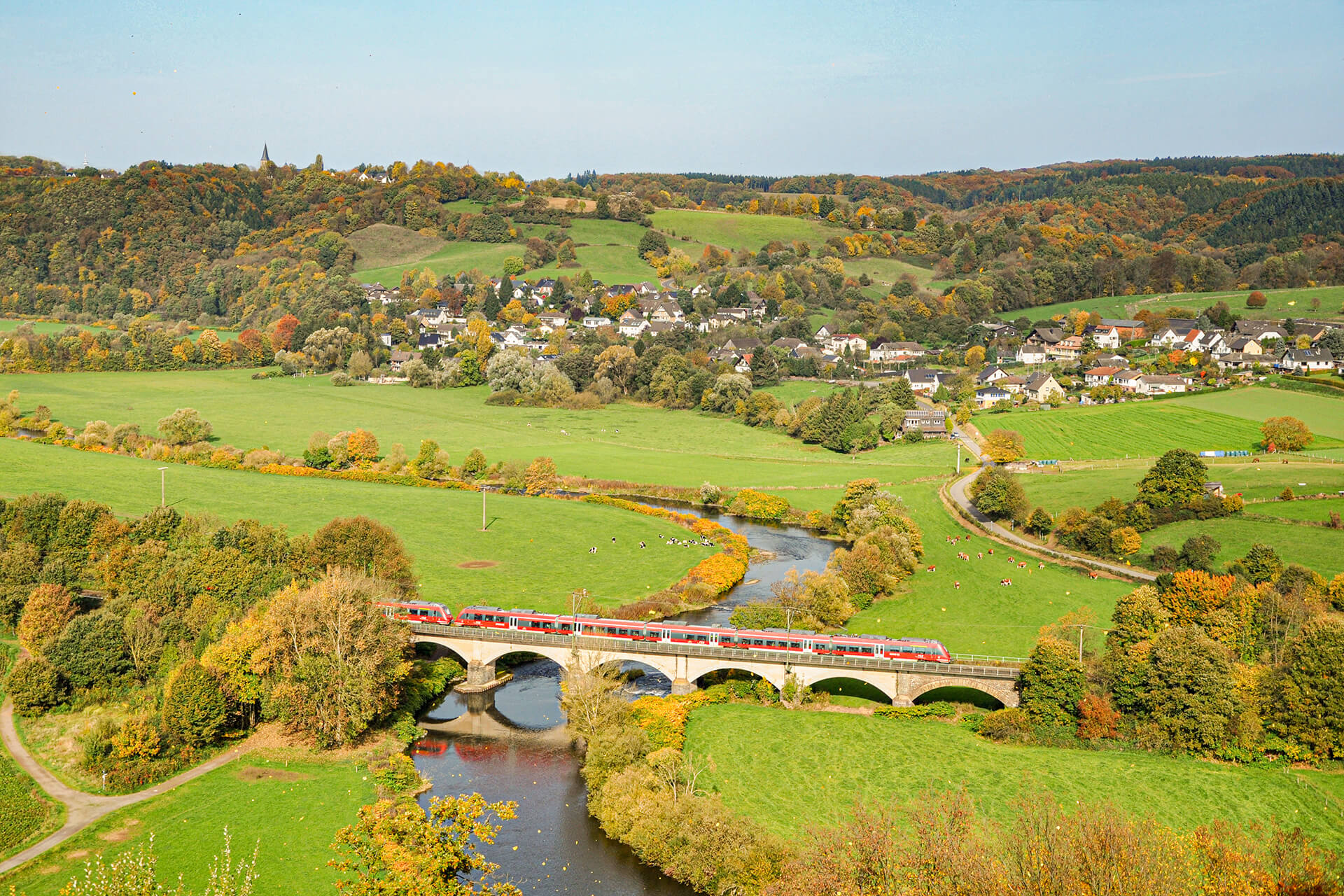 Red DB Regio train crossing a bridge over a river in the countryside with a small town in the background