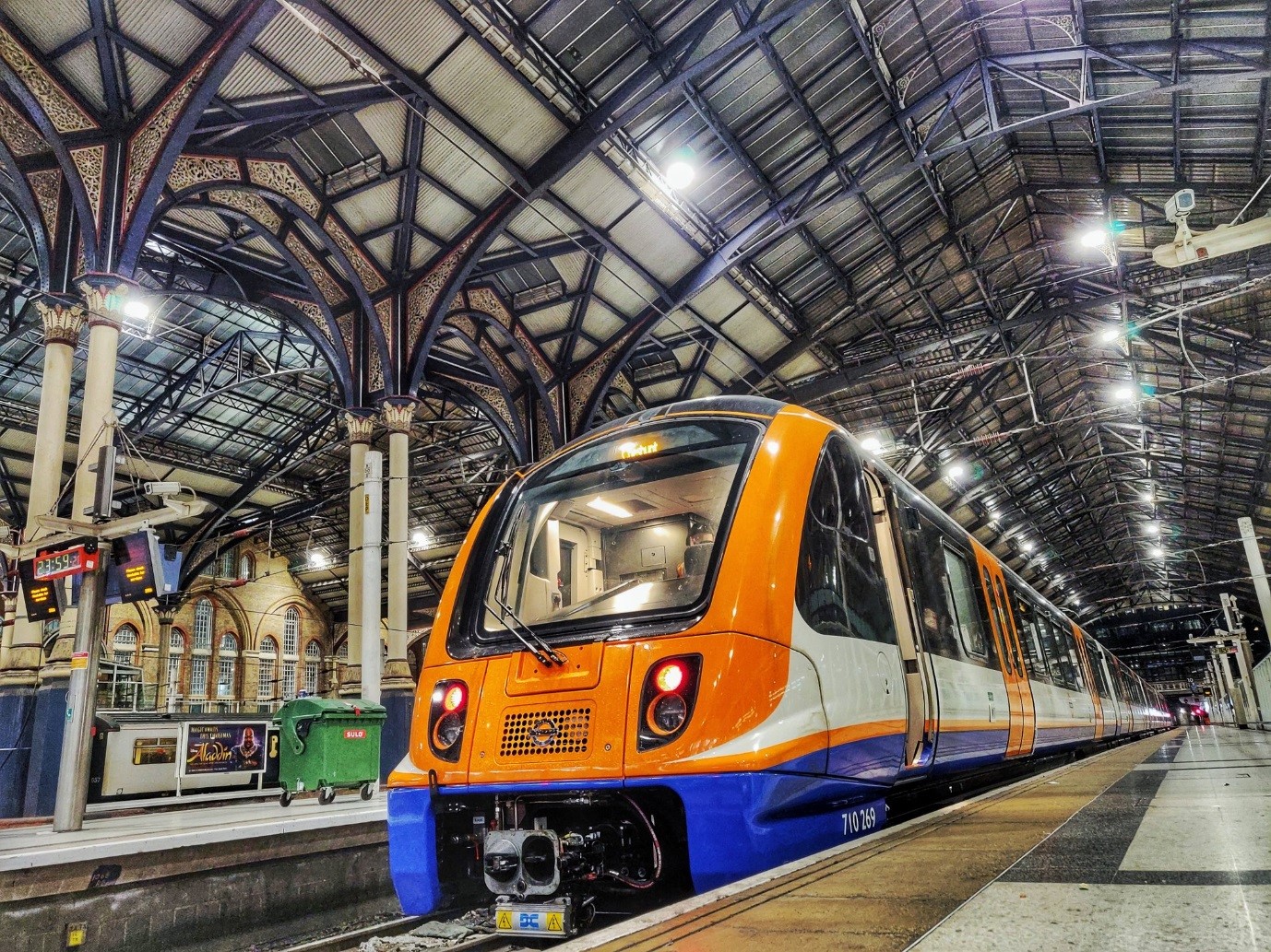 Arriva Rail London train in orange and blue livery at platform inside a covered railway station with glass roof.
