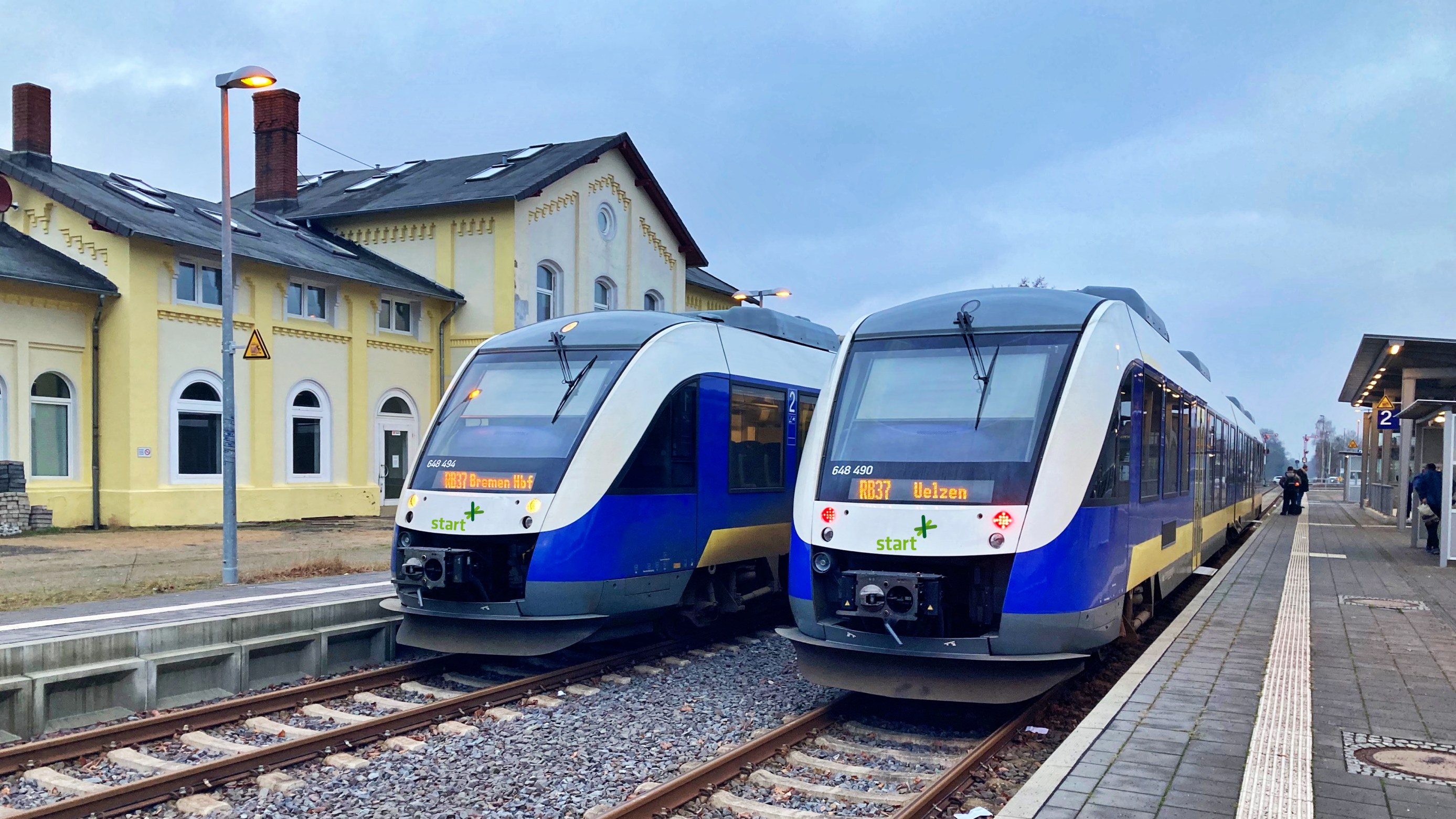 Two Start Niedersachsen Mitte railcars in blue and yellow livery at the platform of a small rural station.