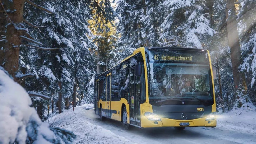 STI Bus AG bus in yellow livery driving through a snowy winter landscape with fir trees.