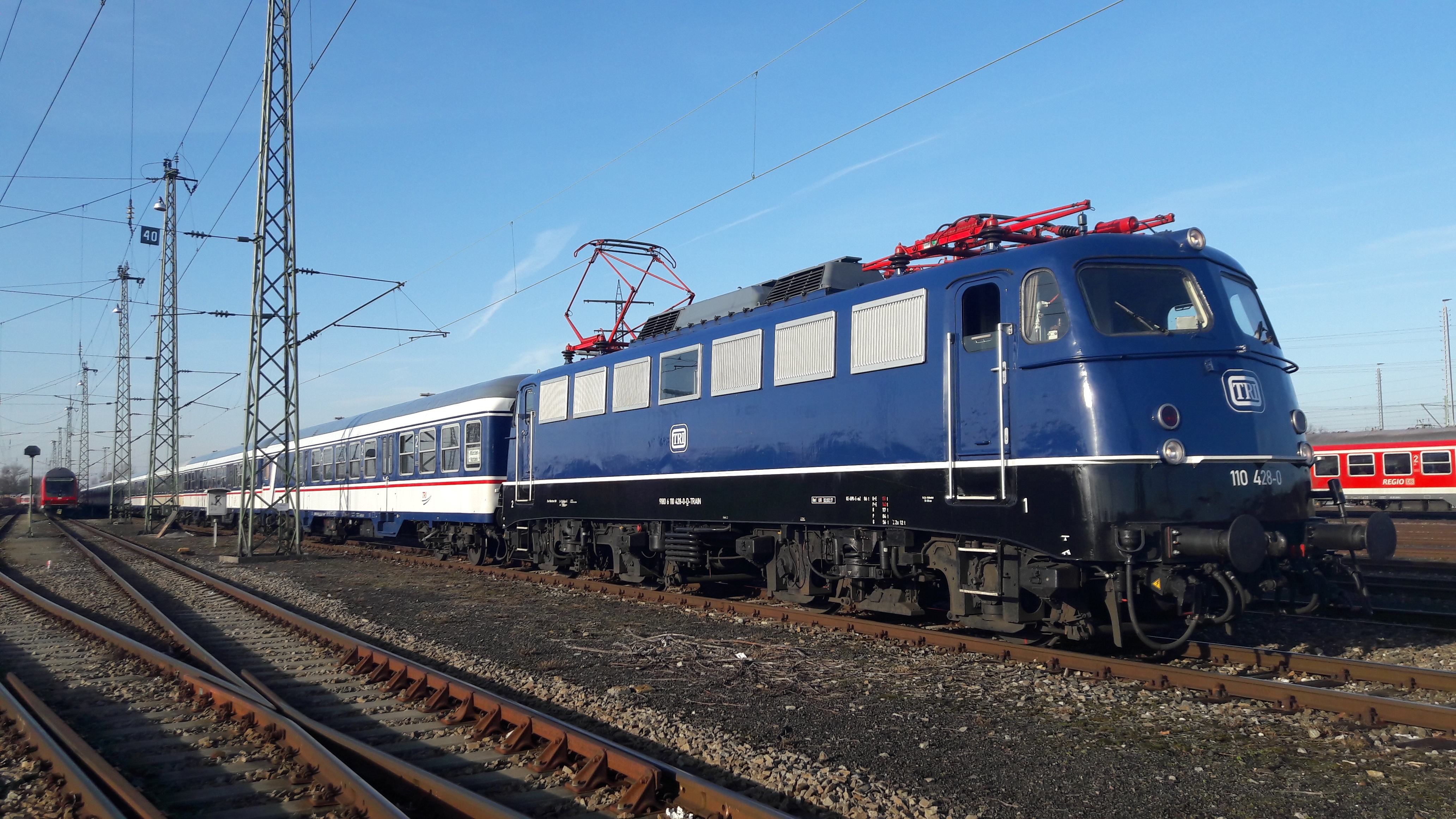 TRI locomotive in blue livery with passenger coaches at a railway yard under clear blue sky.