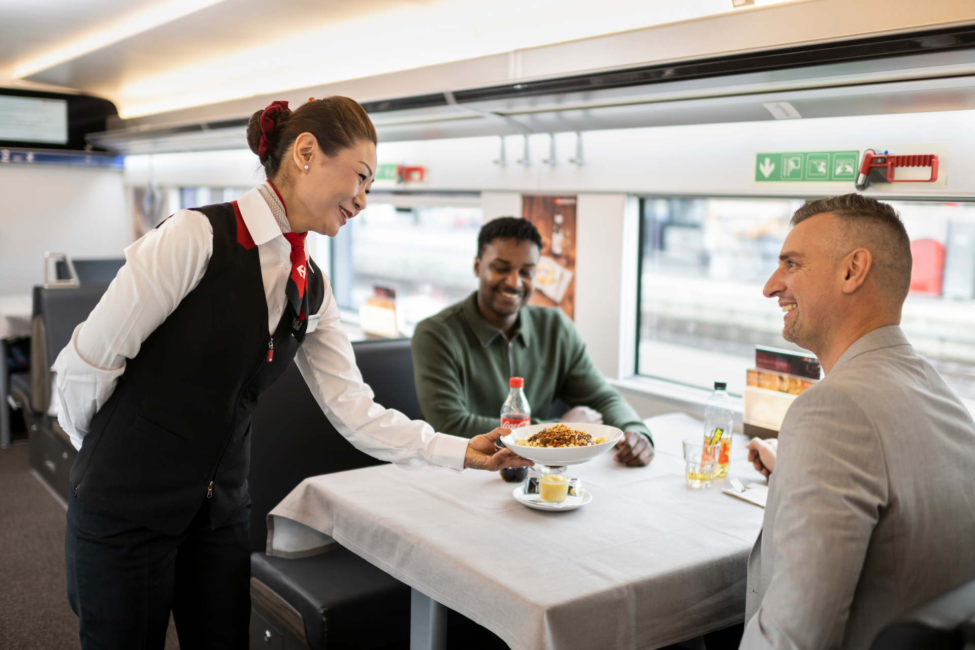 Azafata de tren sirviendo comida a dos pasajeros en mesa del compartimento