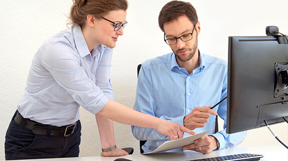 Man discussing with a woman while sitting in front of a computer with a document in his hand